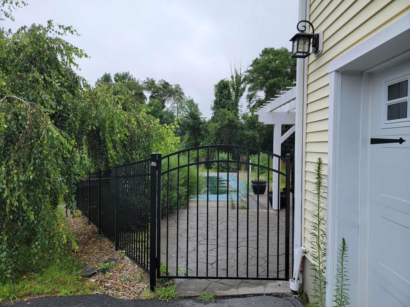 Arched black aluminum pool gate and matching fence beside a yellow house with a lantern sconce and white pergola, opening onto a stamped-concrete patio and in-ground pool framed by tall ornamental grasses.