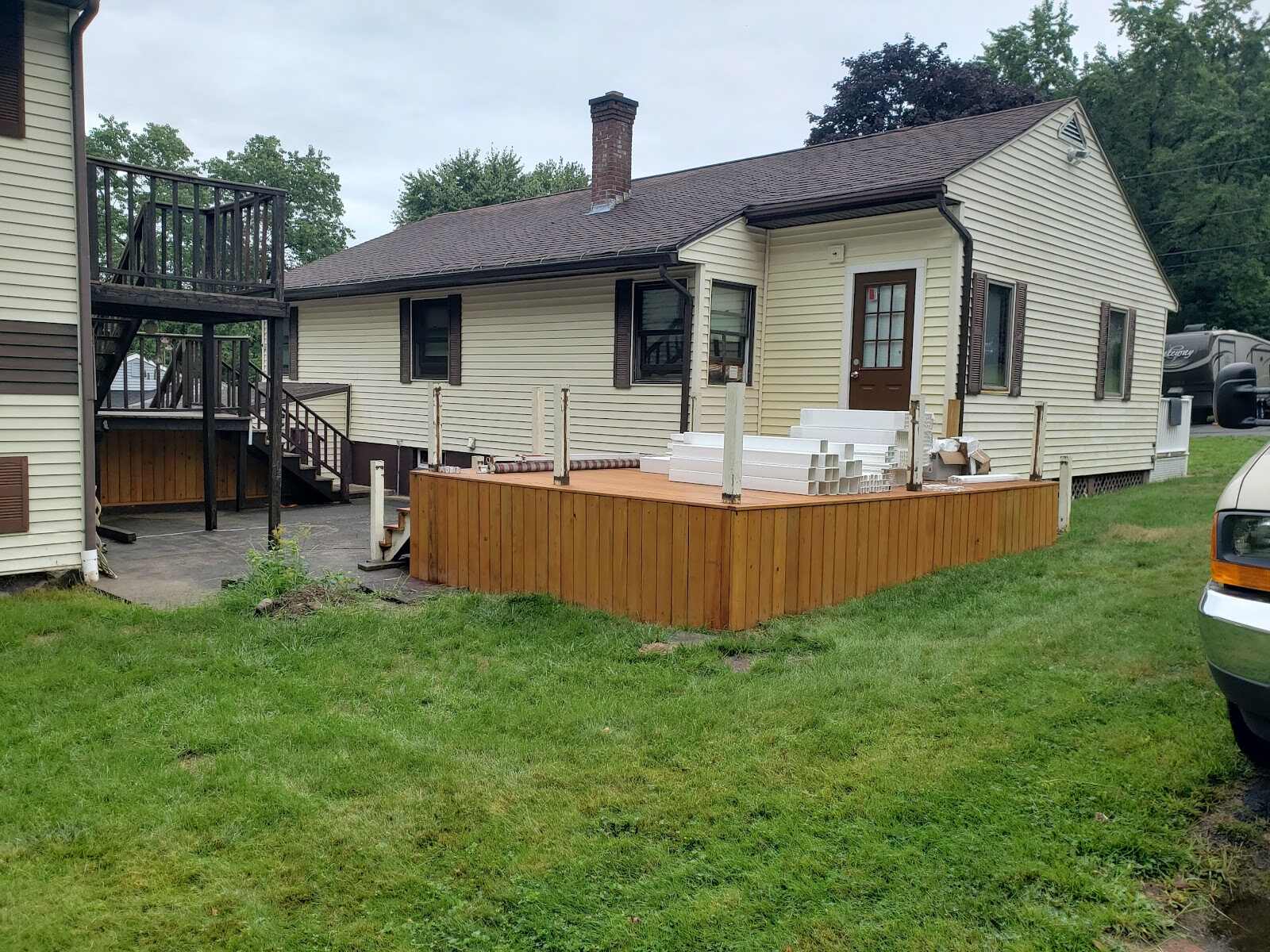 New natural-cedar deck under construction beside a yellow ranch house, with bundled white vinyl railing components and post sleeves stacked on the deck surface ready for installation.