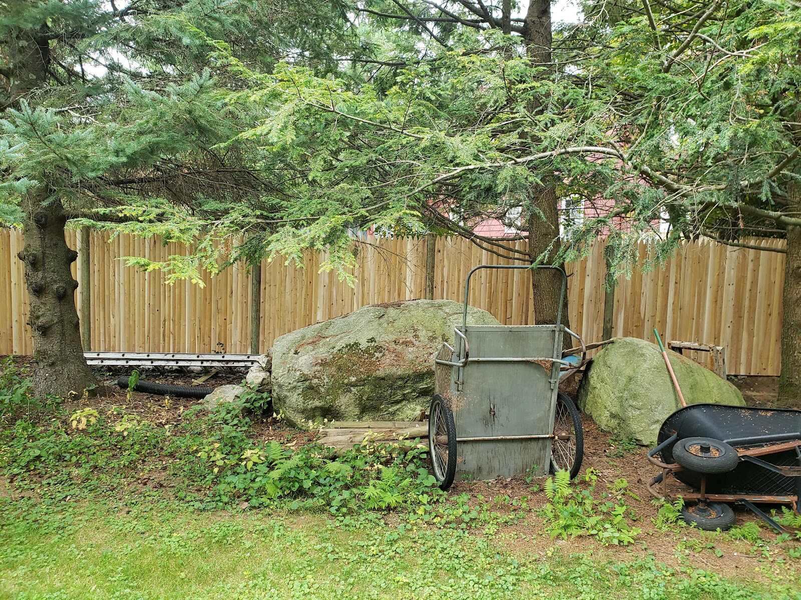 New natural-cedar stockade fence corner in a wooded backyard storage area, with a large boulder, an old two-wheeled garden cart, and scattered yard equipment in front of it.