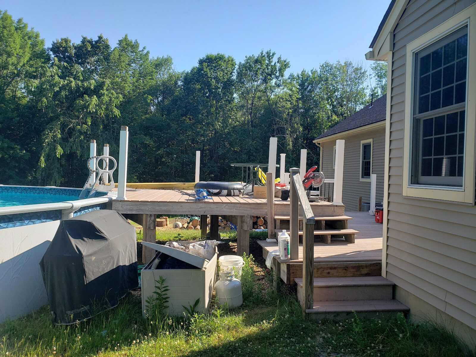 Composite deck under construction with white vinyl railing posts installed, a miter saw and tools on the decking, situated beside an above-ground pool with a slide, next to a tan house.