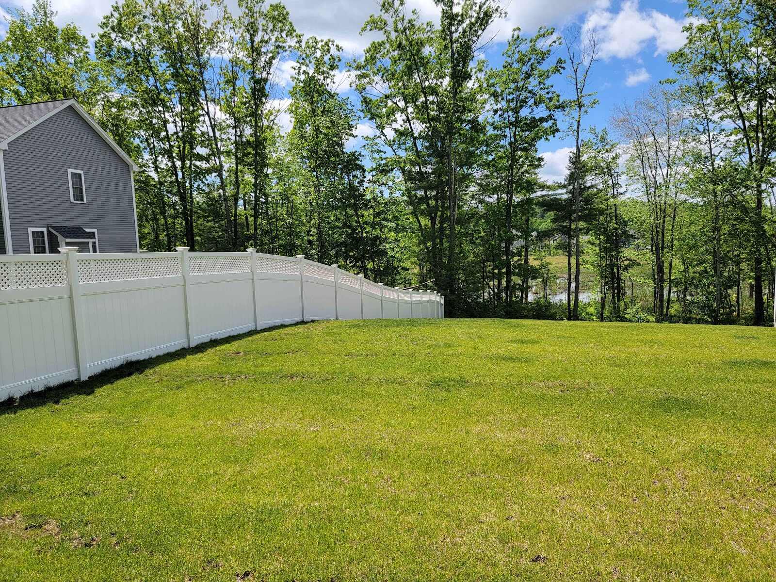 Curved white vinyl privacy fence with a diamond-lattice top following the contour of a sloped, freshly mowed lawn beside a gray house, with a pond visible through the trees beyond.