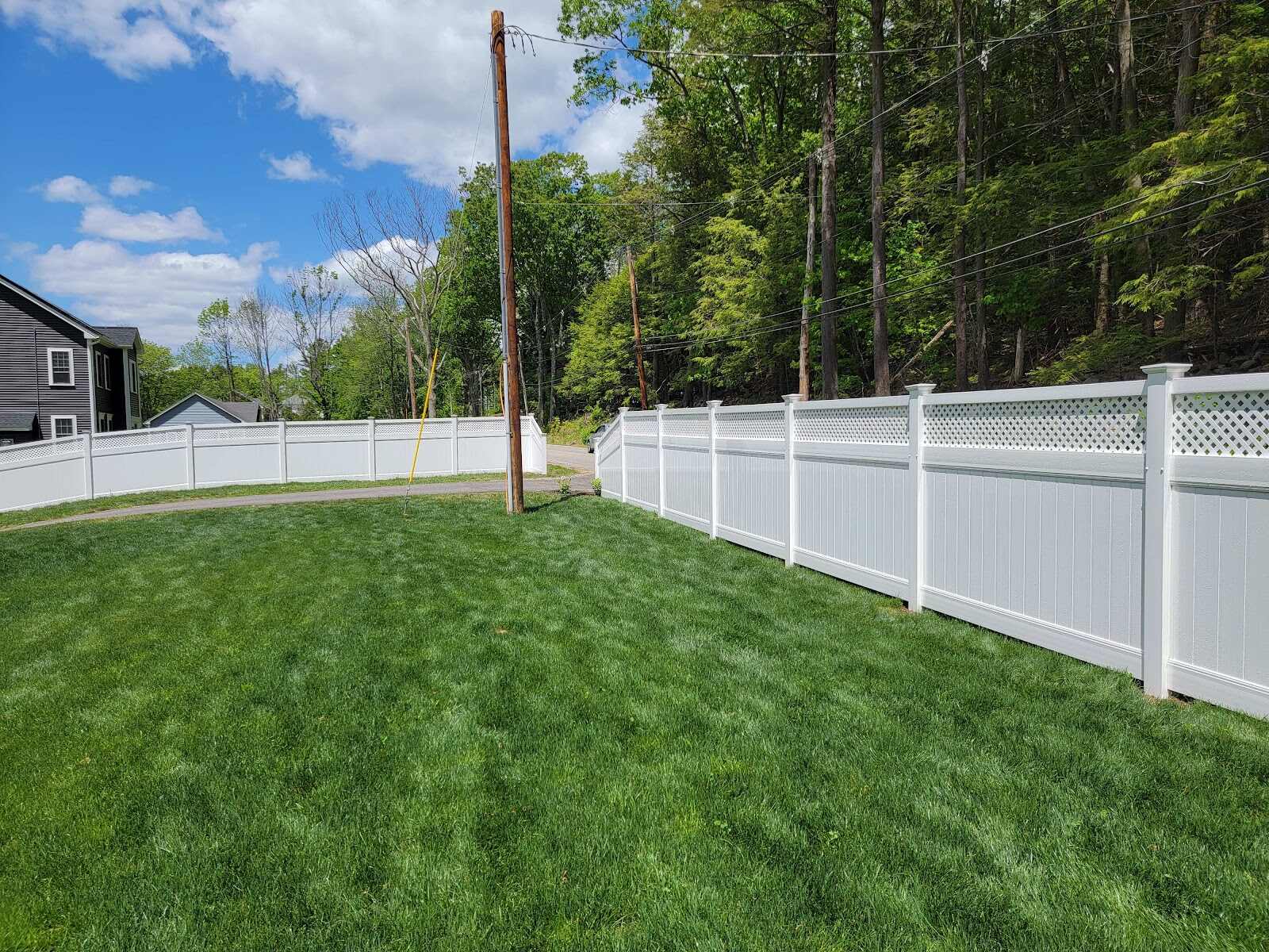 Long, gently curving white vinyl privacy fence with a diamond-lattice top bordering a vividly green, manicured lawn against a bright blue sky with scattered clouds, a gray house and utility pole visible at the fence line.