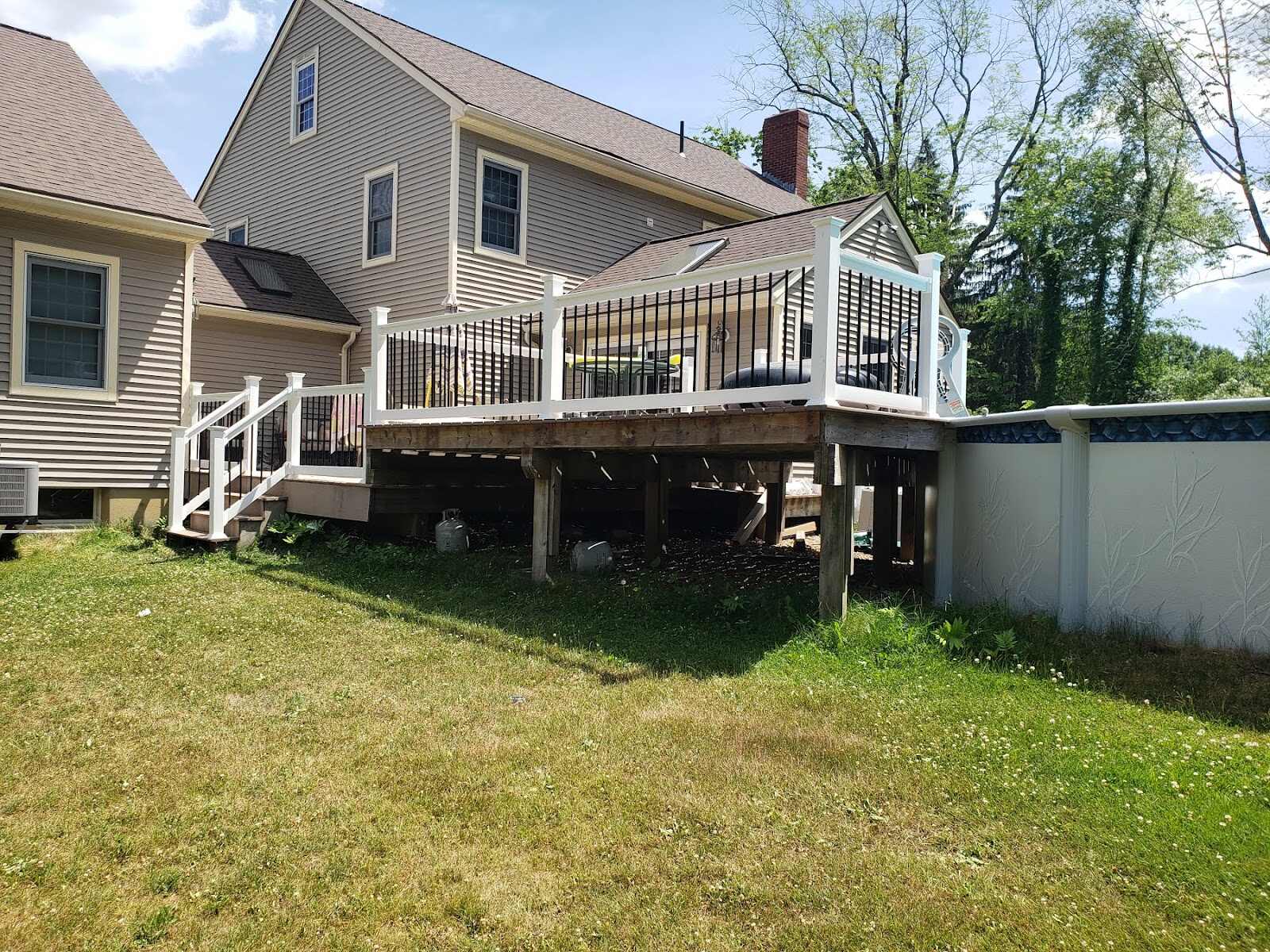 Elevated wood deck with white vinyl posts and black metal picket railing wrapping the rear of a tan two-story house, positioned beside an above-ground pool with a vinyl liner wall.