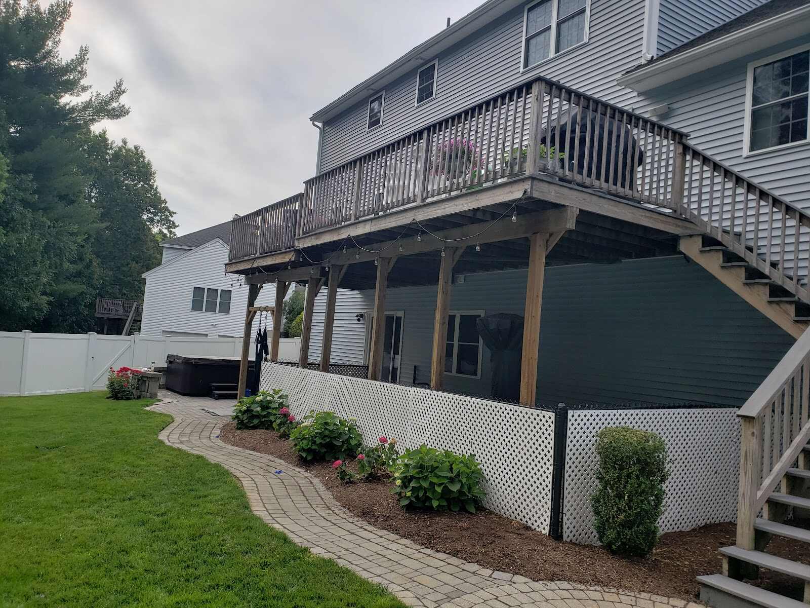 Rear view of a gray two-story house with a large elevated wood deck, a hot tub tucked beneath it behind white vinyl lattice skirting, a curving paver walkway through mulched planting beds, and a white vinyl privacy fence at the property line.