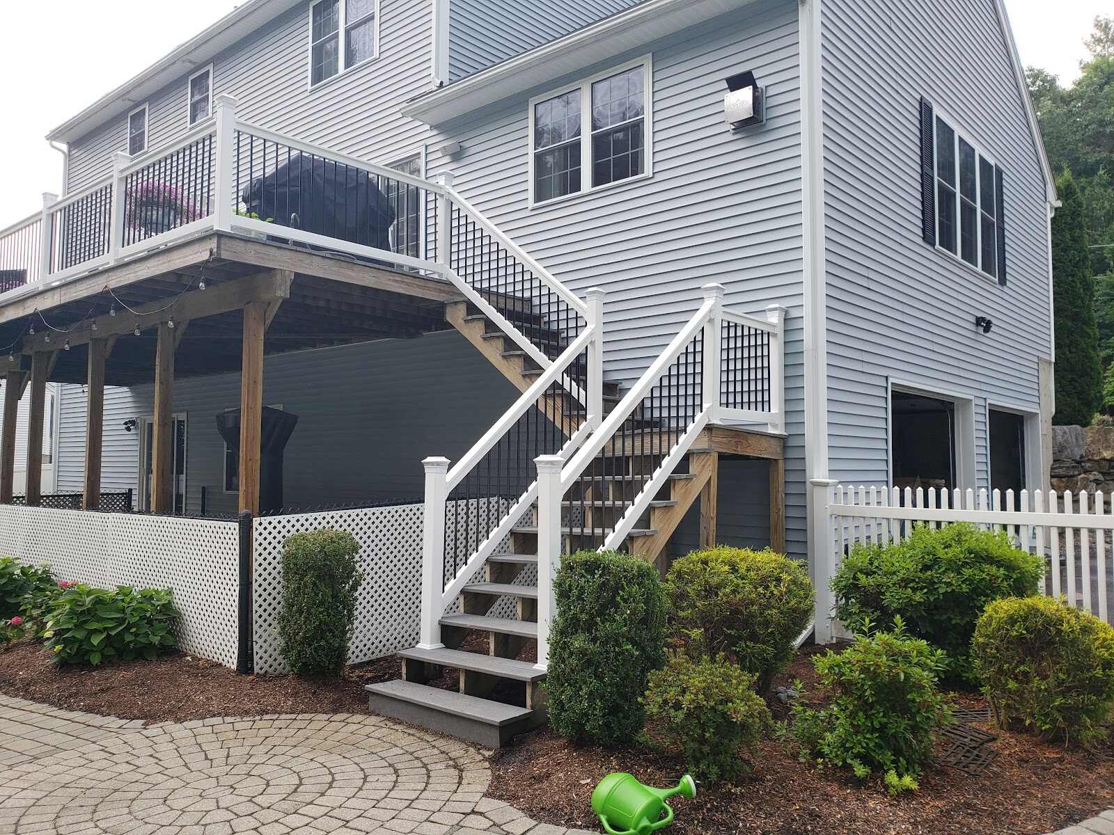White vinyl and black metal picket deck staircase descending from an elevated deck to a paver patio, alongside white vinyl lattice skirting and a white picket fence at a blue-gray house.