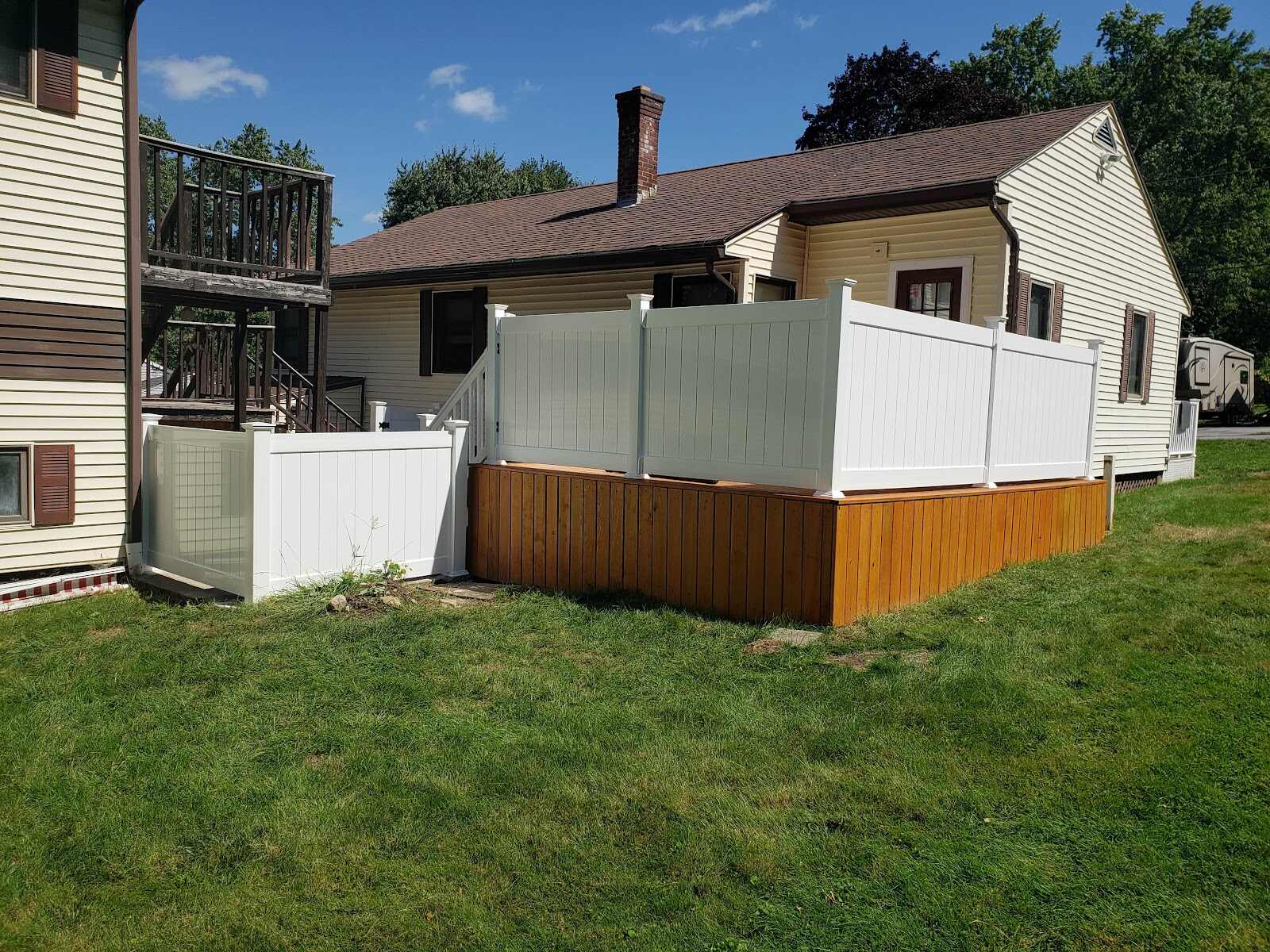 Completed white vinyl privacy fence with a black-picket accent top installed atop a natural-cedar deck skirt wall and matching low fence section, wrapping the corner of a yellow ranch house with a shed alongside.