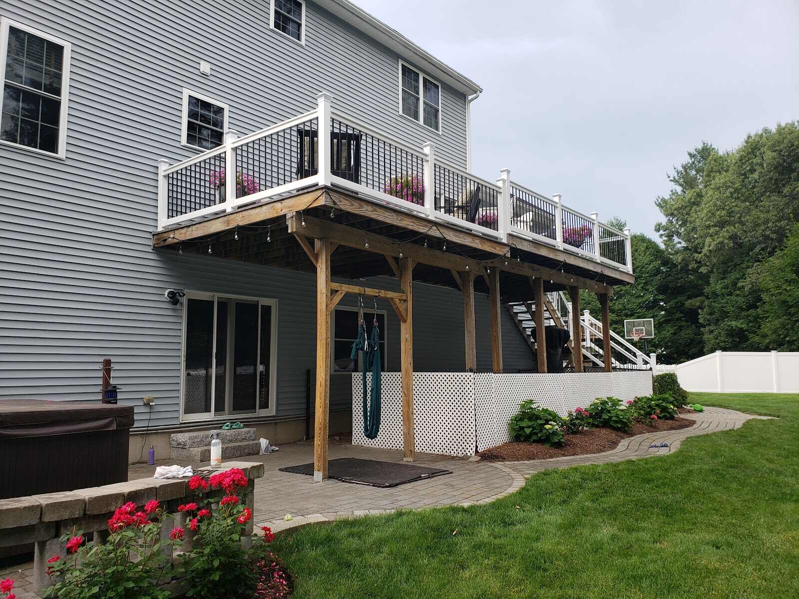 Elevated wood deck with white vinyl lattice skirting beneath it, string lights, potted flowers on the railing above, and a white vinyl privacy fence visible along the property line behind a blue-gray house with a hot tub and paver patio.