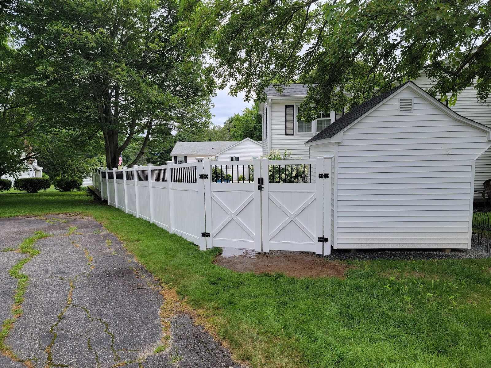 Long run of white vinyl privacy fence with a black-picket accent top and a wide double X-brace gate, running from a cracked driveway past a white storage shed toward a two-story house under mature shade trees.