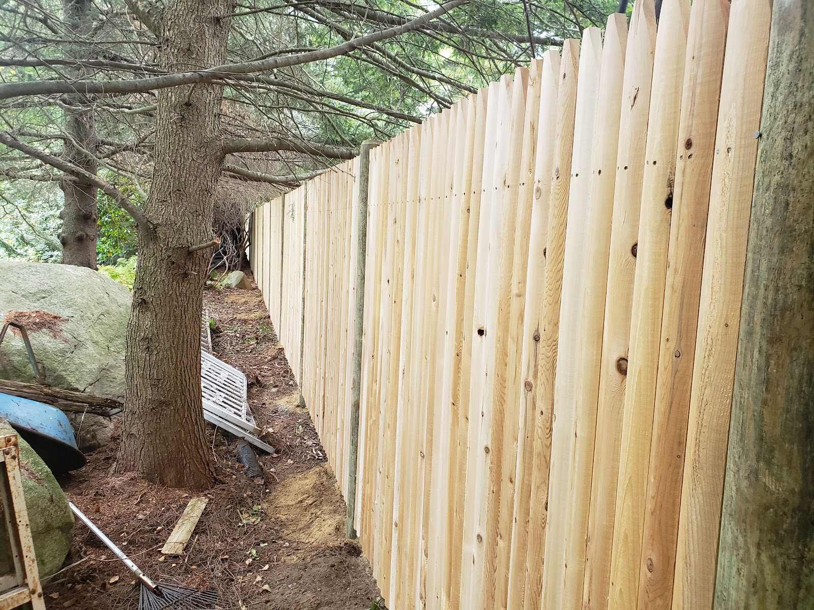 Close-up of a freshly built natural-cedar stockade (dog-ear picket) fence running past a large tree trunk, with a boulder, tarps, and yard debris piled against it on the wooded side.