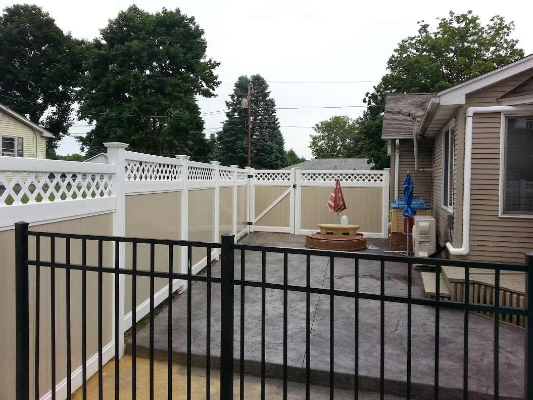 Backyard concrete patio bordered by a black aluminum picket fence in the foreground and a tan/white vinyl privacy fence with a matching gate behind, a small tiered fountain, red patio umbrella, and above-ground pool equipment against a tan ranch house.