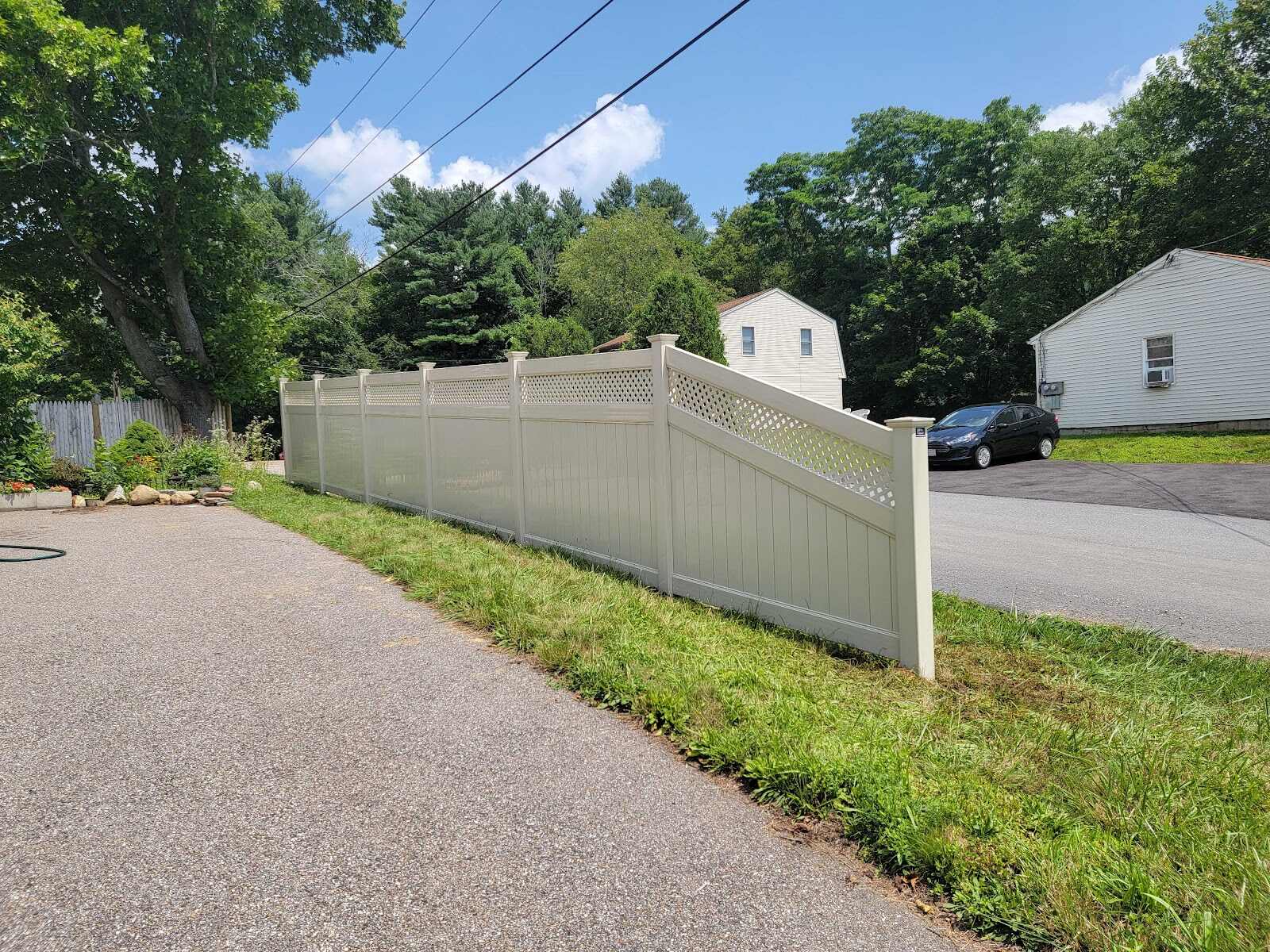 Tan/almond vinyl privacy fence with a diamond-lattice top section stepping down a grassy slope beside a paved driveway, with houses and a parked car visible across the street under a bright blue sky.