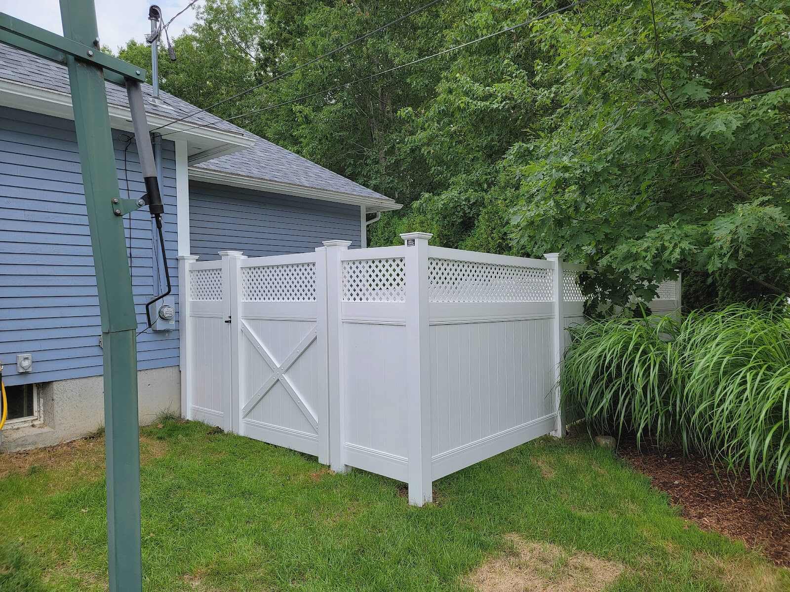 White vinyl privacy fence corner with a diamond-lattice top and an X-brace gate screening a utility meter area beside a blue house, bordered by ornamental grasses.