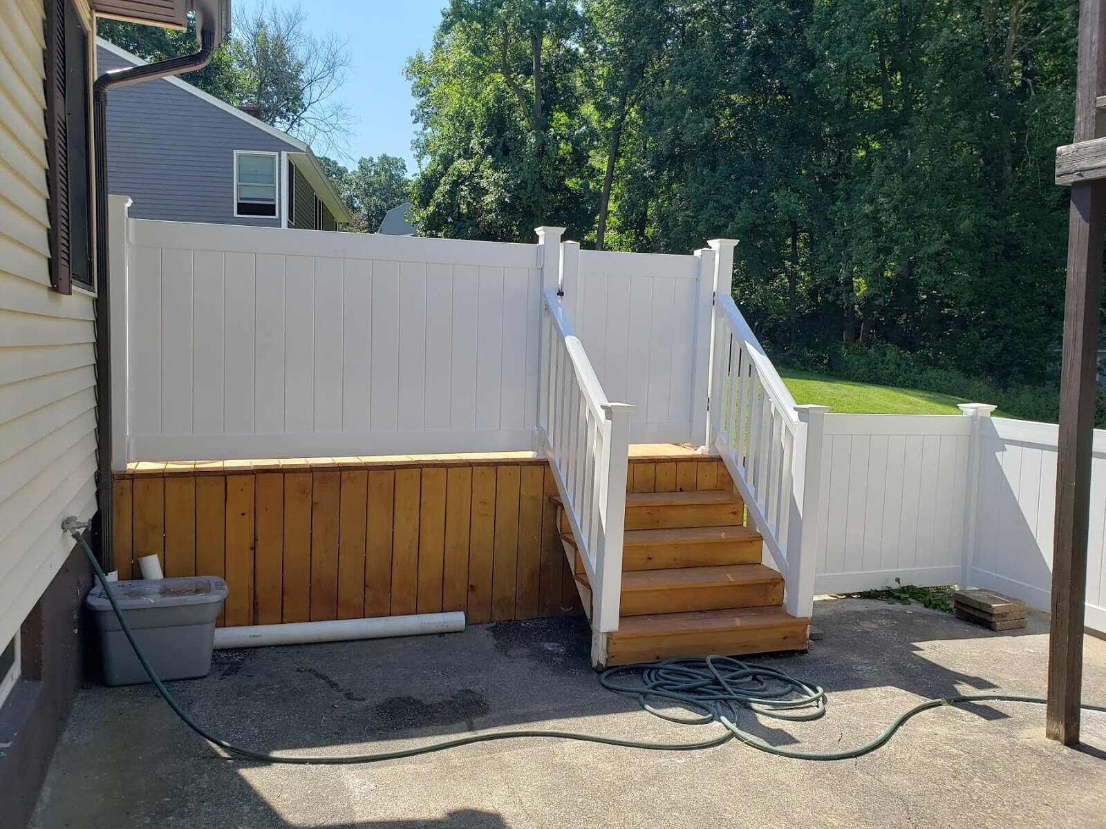 White vinyl privacy fence and gate atop a natural-cedar deck skirt wall, with a matching staircase leading down to a concrete patio beside a yellow house, wooded lot behind.
