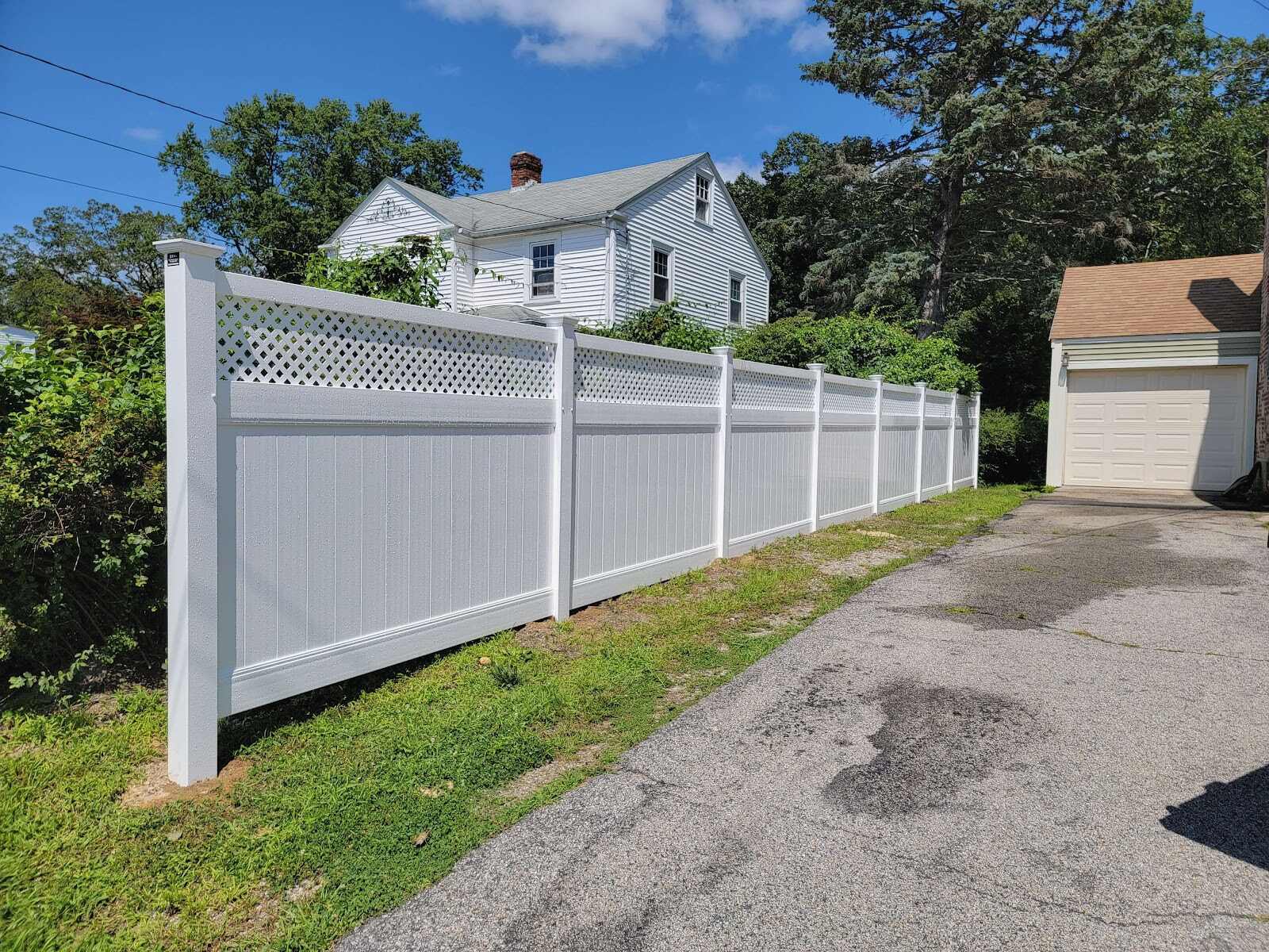 White vinyl privacy fence with a diamond-lattice top running beside a paved driveway toward a detached garage, a white farmhouse with a brick chimney visible above the fence line under a clear blue sky.