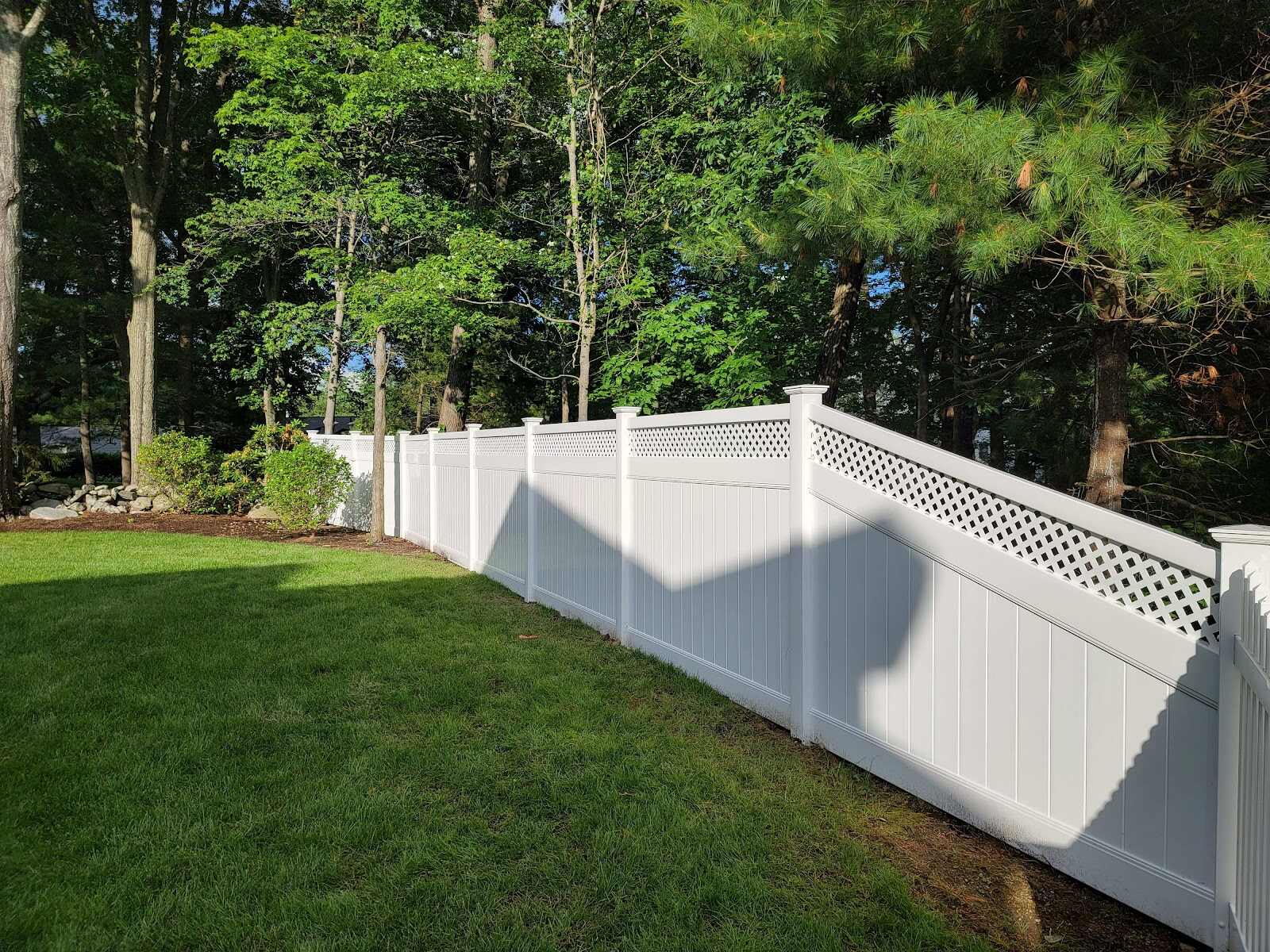 White vinyl privacy fence with diamond-lattice top panel running along a lush green lawn bordered by a stone edging and dense pine/maple woods, evening sun casting long shadows.