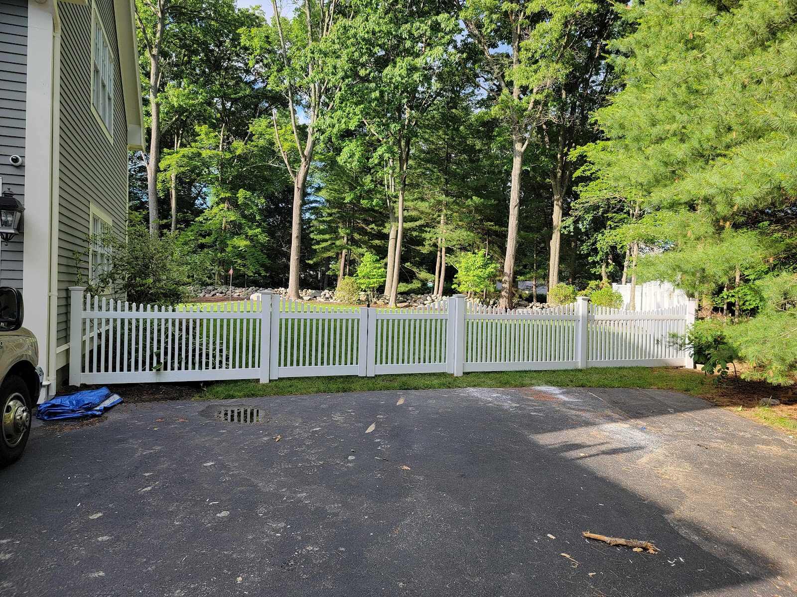 White vinyl picket fence with a scalloped top rail running along a paved driveway beside a house, backed by a dense stand of tall mature trees.