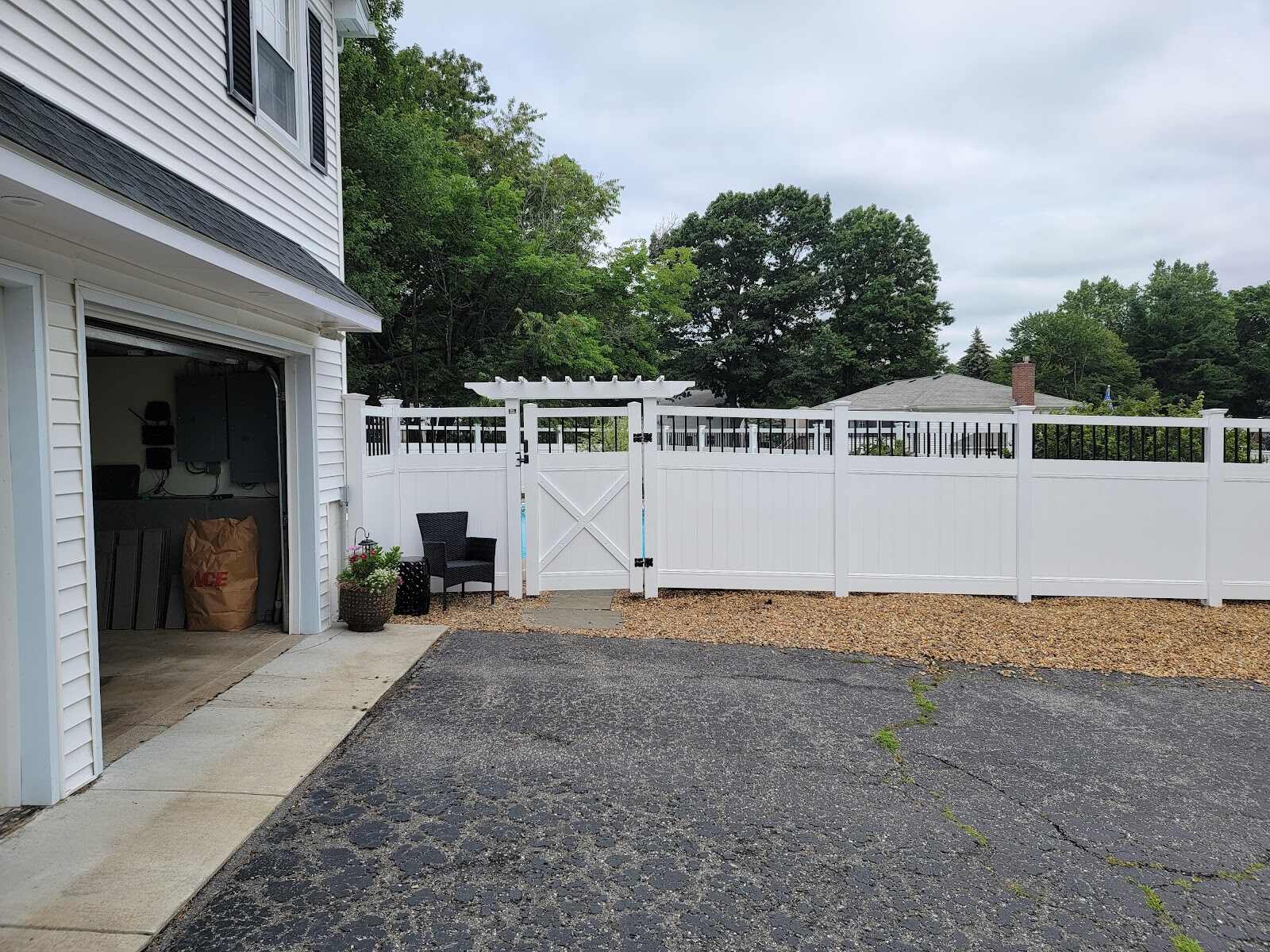 White vinyl privacy fence with a black-picket accent top and a matching X-brace gate topped by a white lattice arbor, opening onto a gravel-bordered driveway with a black wicker chair, leading toward an in-ground pool.