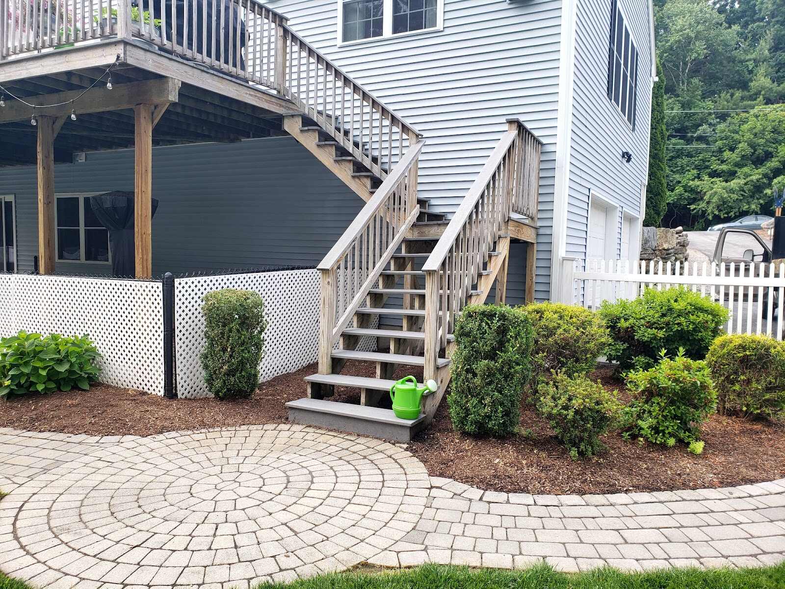 Weathered wood deck staircase with white vinyl railing descending past white vinyl lattice skirting to a circular paver patio, boxwood shrubs and a green watering can at the base, a white picket fence and parked truck visible beyond.